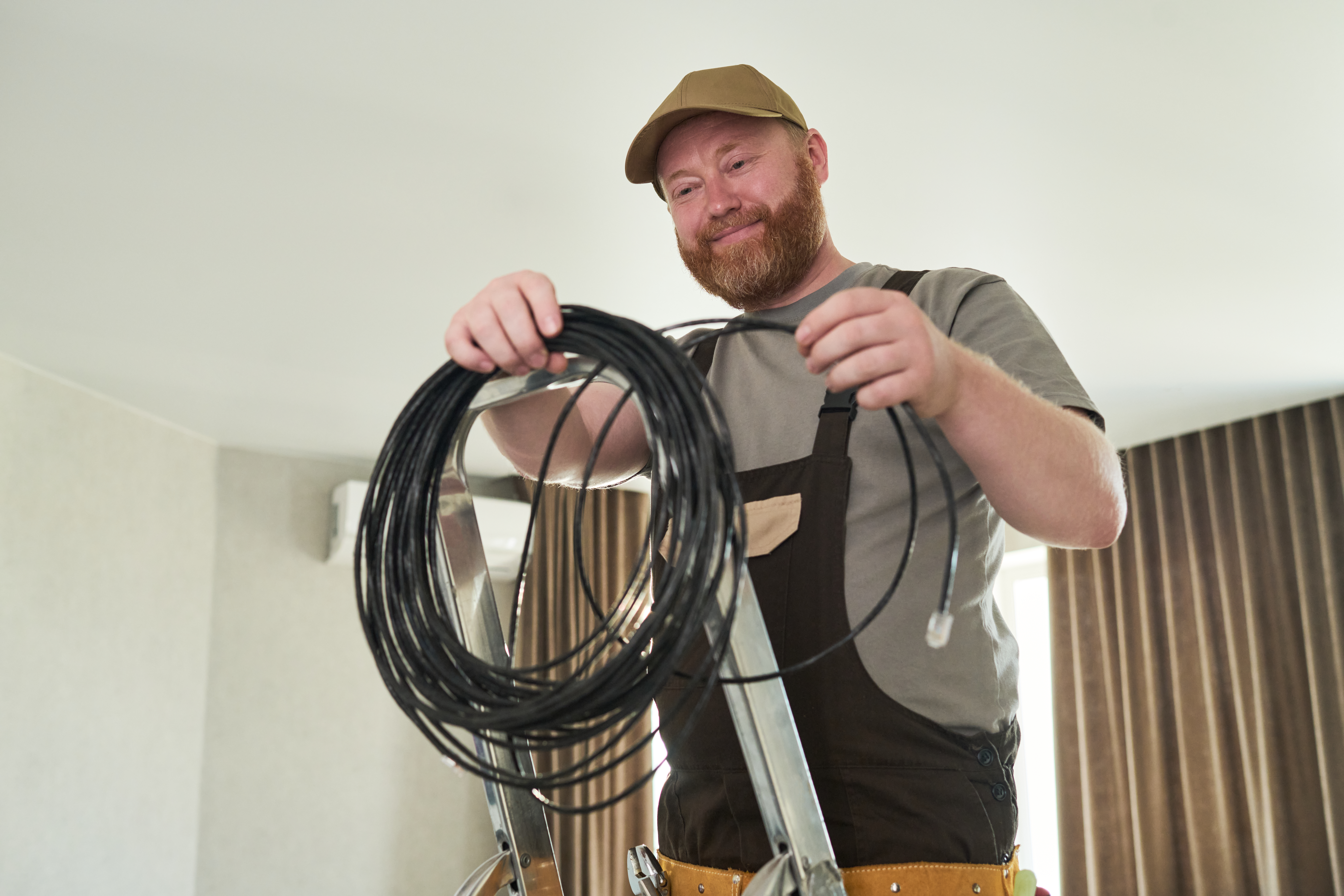 Caucasian middle aged man standing on ladder holding coiled cable smiling while preparing for electrical installation in modern room with curtains visible in background